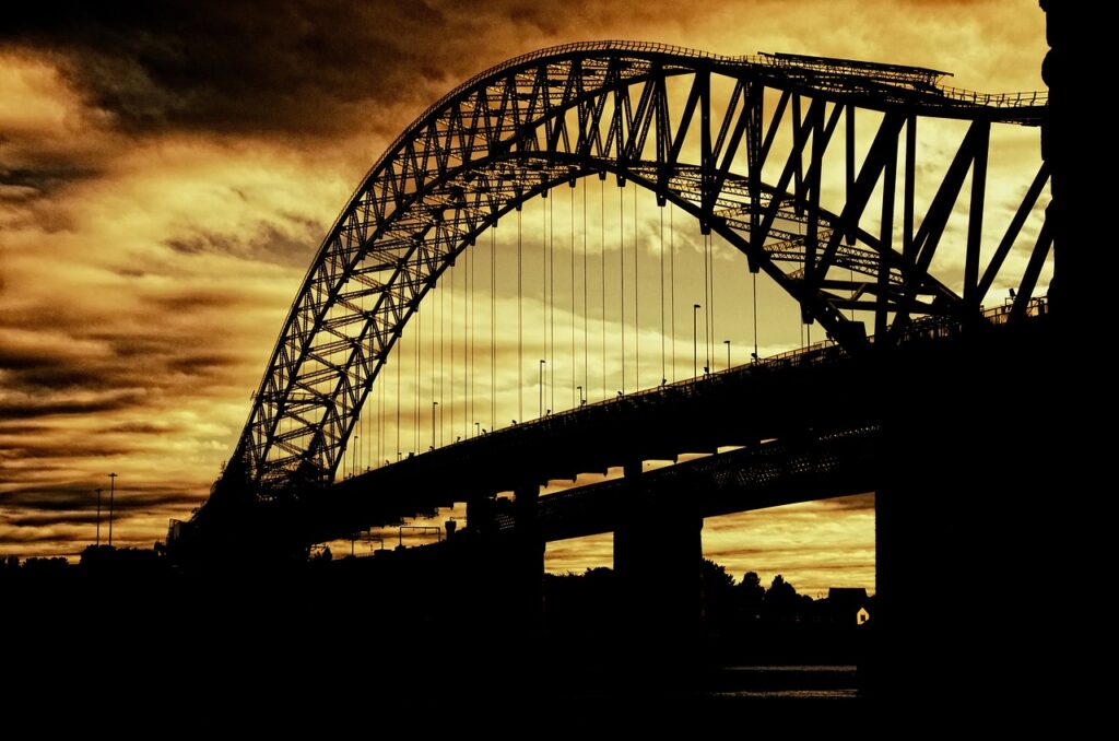 image d'un pont en métal et pierre, de style arche moderne. La photo a été prise au moment du crépuscule, laissant paraitre le pont tel une ombre